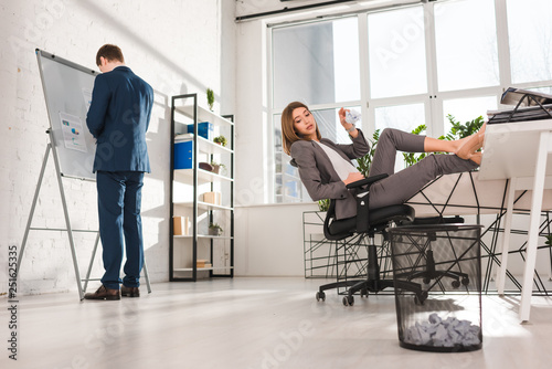 selective focus of attractive businesswoman sitting on chair and throwing paper in bin near coworker, procrastination concept photo