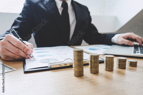 Businessman writing making report with stacked coins arranged at office desk and many document data graph, business concept