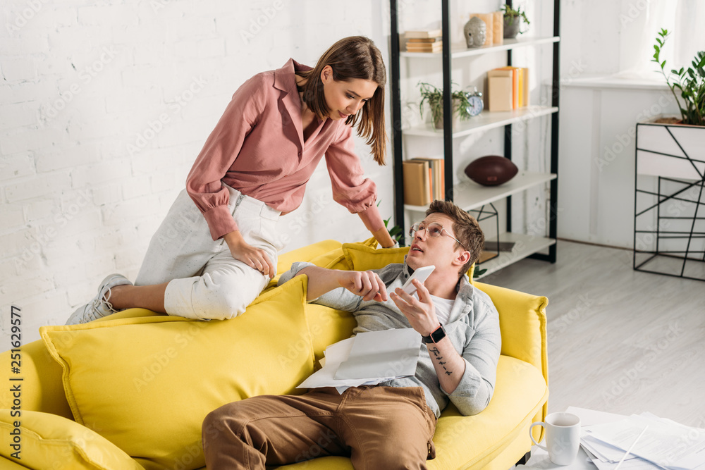 attractive woman looking at boyfriend with smartphone lying on sofa attractive woman looking at boyfriend with smartphone lying on sofa
