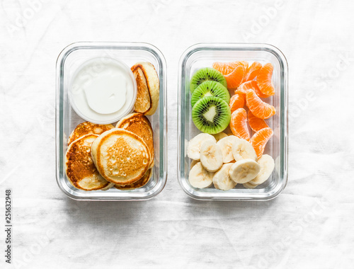 Healthy kids school lunch box - pancakes with sour cream and banana, kiwi, tangerine fruit. Delicious snack on a light background, top view