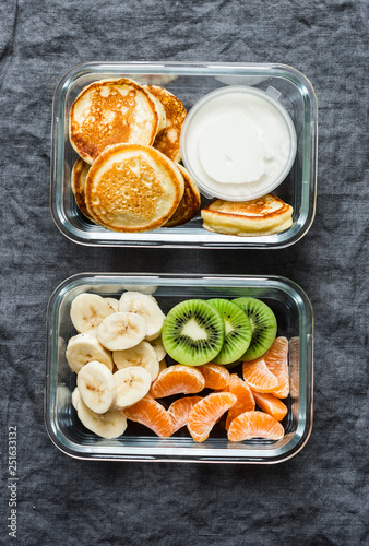 Healthy kids school  walk  picnic lunch box - pancakes with sour cream and banana  kiwi  tangerine fruit. Delicious snack on a grey background  top view