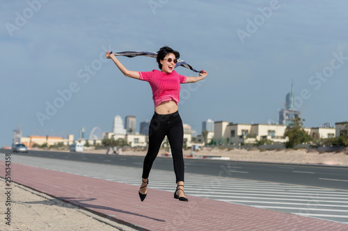 Beautiful European caucasian woman jumping and laughing on the side of a city highway wearing pink shirt and black tight pant and a playing with her scarf 