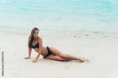 Sexy model in black swimsuit lying on white sandy beach. Girl sunbathes and rests on vacation at tropical island