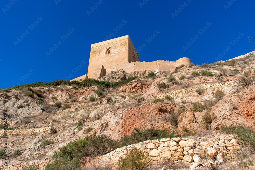 A steep rocky hill leading up to the square tower of Lorca Castle framed by an ultra blue sky in southern Spain