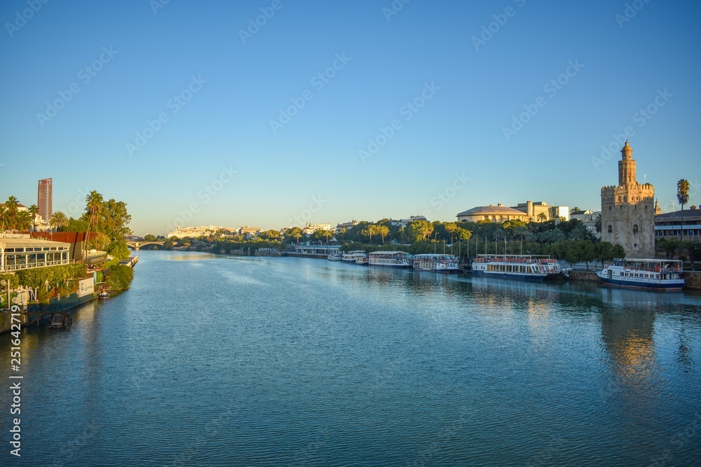 The Guadalquivir river that flows through Seville in south Spain with riverboat cruise boats lining the riverfront in the early morning, with the rising sun just touching the Golden Tower.