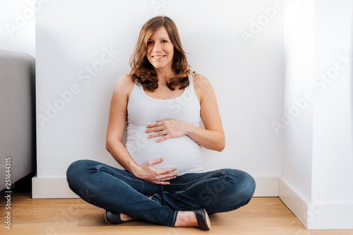 joyful pregnant woman in white top sitting on the ground holding her belly