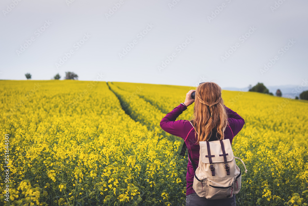 Hiking woman looking through binoculars. Bird watching in blooming field Hiking woman looking through binoculars. Bird watching in blooming field