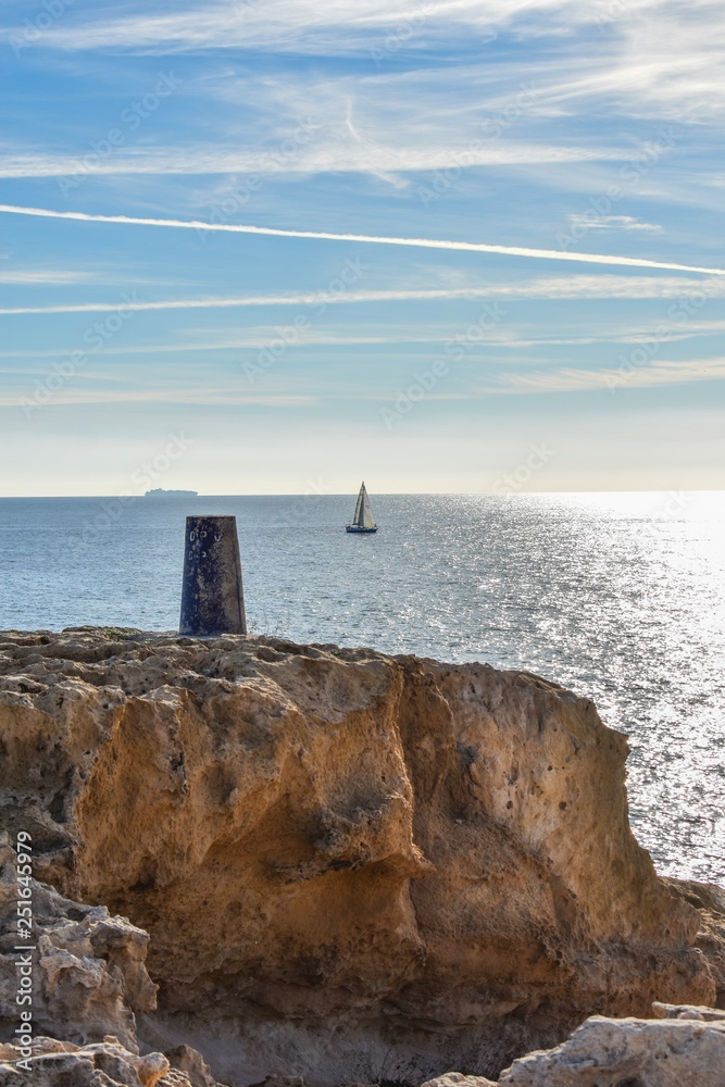 A location marker situated along the rocky coastline of southern Spain looks over the tranquil water of the Mediterranean where a small sailboat sails past and a much larger ship sits on top of the ho