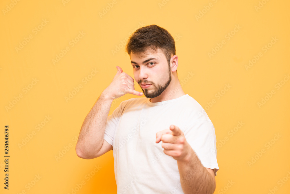 Man with a beard with a smile looks at the camera and shows a hand gesture of the phone, call me. Man in a white T-shirt shows a finger in the camera , isolated on a yellow background. Man with a beard with a smile looks at the camera and shows a hand gesture of the phone, call me. Man in a white T-shirt shows a finger in the camera , isolated on a yellow background.