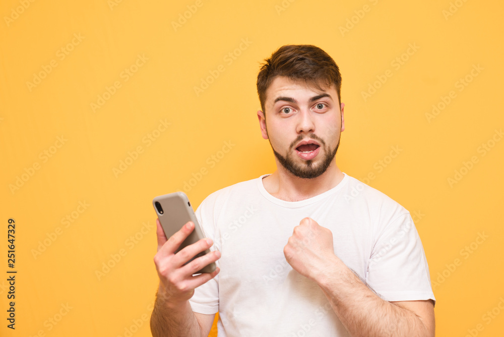 Emotional man with a beard, wearing a white T-shirt, stands on a yellow background with a smartphone in his hands and looks surprised at the camera. Isolated. Copyspace Emotional man with a beard, wearing a white T-shirt, stands on a yellow background with a smartphone in his hands and looks surprised at the camera. Isolated. Copyspace