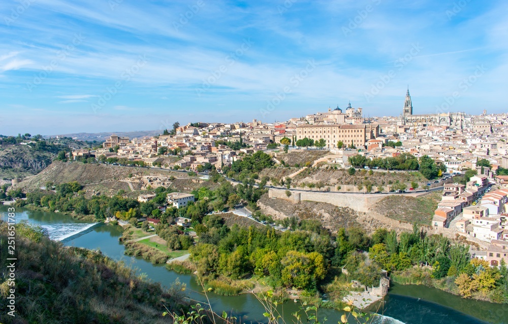 A panoramic view of the old city of Toledo, with it’s defensive walls and the tower of the cathedral inside the city walls, surrounded by a ravine and river that serves as a moat.