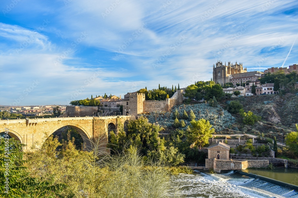 A view of an ancient bridge over the river moat of Toledo, Spain, spanning 3 arches and an old fortified gatehouse, with houses on the far side creeping up the hill and lush green trees at the bases o