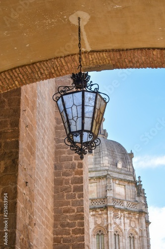 An ornate cast iron lamp with frosted glass hangs from the bottom of a bridge with the tower of Toledo’s cathedral coming into view