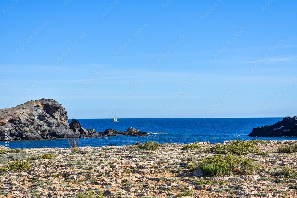 The dry semi-desert of southern Spain meets the blue water of the mediterranean with rocks rising above the water and a small sailboat sailing, along the coast.