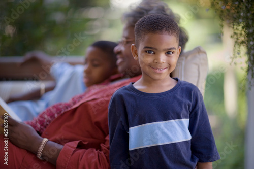 Portrait of a young boy standing beside his mature grandmother lying on a couch reading with his brother. photo