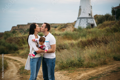 Happy young family in white t-shirts and blue jeans with a small daughter near to the white lighthouse, outdoors background