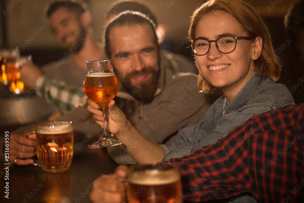Pretty red haired woman with ginger hair posing in beer pub. Beautiful female client looking at camera and smiling. People in bar holding beer glasses in hand.
