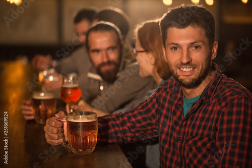 Handsome man in checked shirt holding glass of beer while sitting in bar. Bearded man smiling and looking at camera. Background of people talking and resting in beer pub.