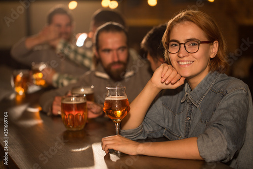 Adorable woman in spectacles posing in beer pub full of clients. Young woman smiling  looking at camera and holding hand near chin. Female client holding glass of delicious beer.