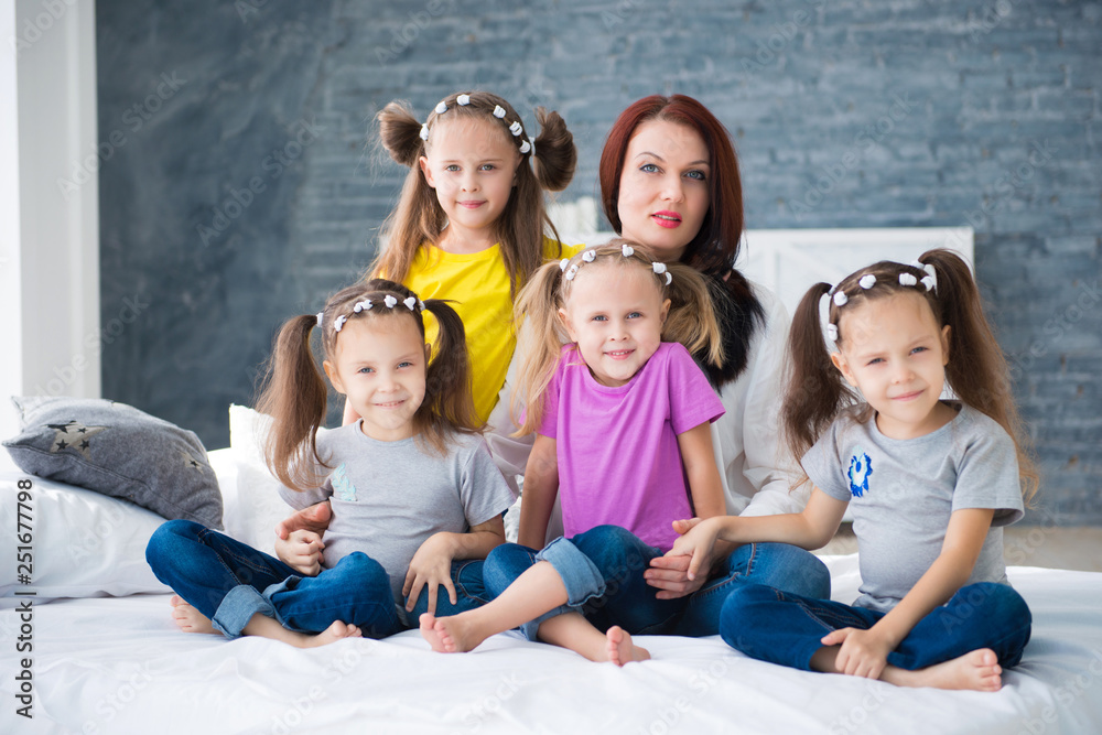Obraz premium Large friendly family, many children: mom and four pretty cheerful girls triple twins sisters sitting on a bed against a gray brick wall