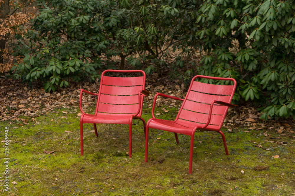 Side view two red chairs in outside green natural park. On green grass with plants around them. Side view two red chairs in outside green natural park. On green grass with plants around them.