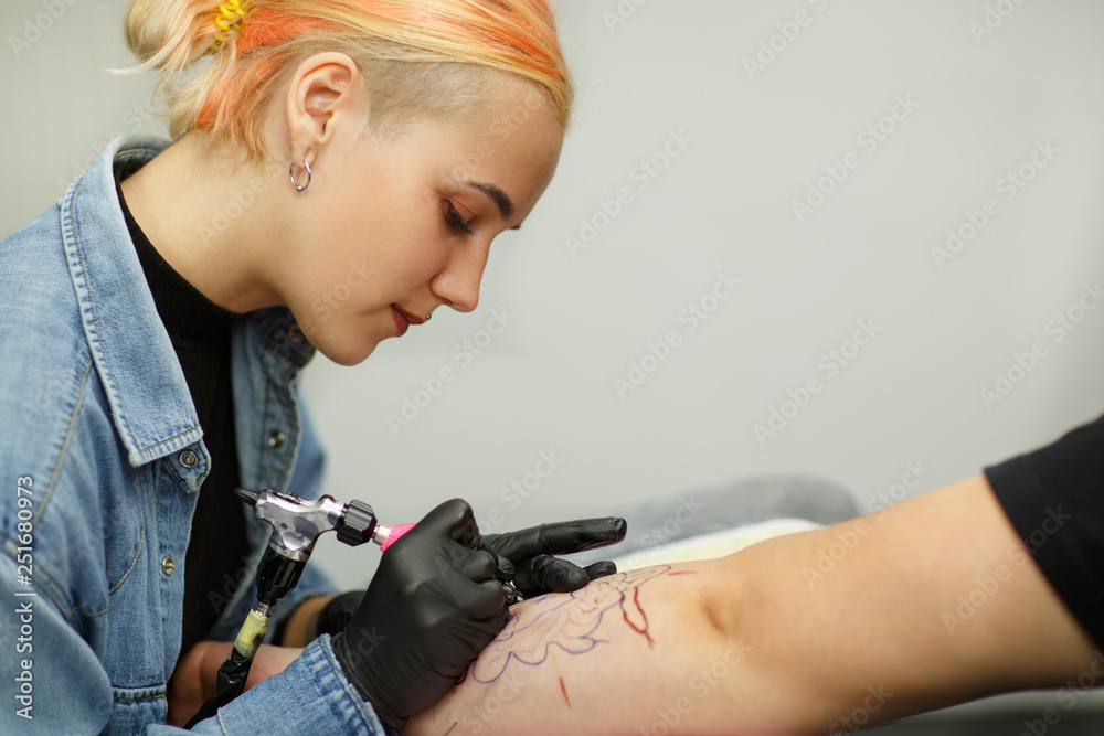 Side view of female tattoo master working and drawing picture on client's hand. Tattooist wearing in black gloves, using tattoo machine and ink. Concentrated and serious girl looking down. Side view of female tattoo master working and drawing picture on client's hand. Tattooist wearing in black gloves, using tattoo machine and ink. Concentrated and serious girl looking down.
