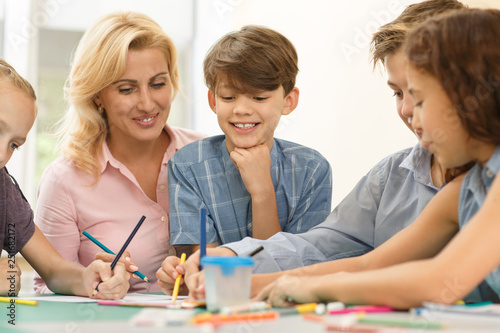 Cute children with teacher sitting at desk and painting on paper.