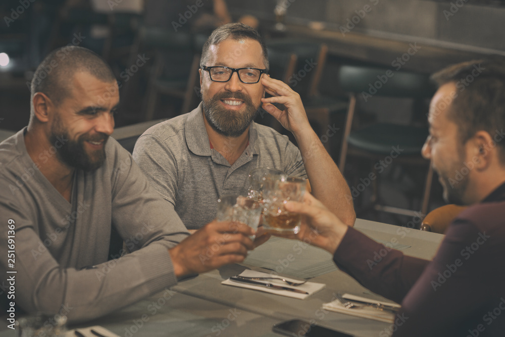 Company having meeting in bar. Men clinking crystal glasses and smiling. Handsome bearded man wearing in spectacles looking at camera. Friends drinking alcoholic beverages, brandy or whisky.