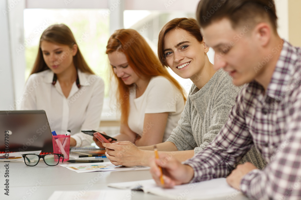 Selective focus of female student looking at camera and smiling while classmates writing and doing test on courses. Pretty girl enjoying interesting studying in modern school. Concept of happiness. Selective focus of female student looking at camera and smiling while classmates writing and doing test on courses. Pretty girl enjoying interesting studying in modern school. Concept of happiness.