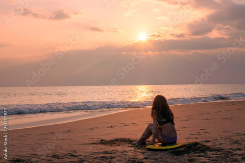 Girl with a surfboard sits on the beach and looks at the sunset © Serge Touch