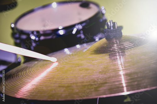 Drumstick playing on a hi-hat or ride cymbal. Drum kit at background