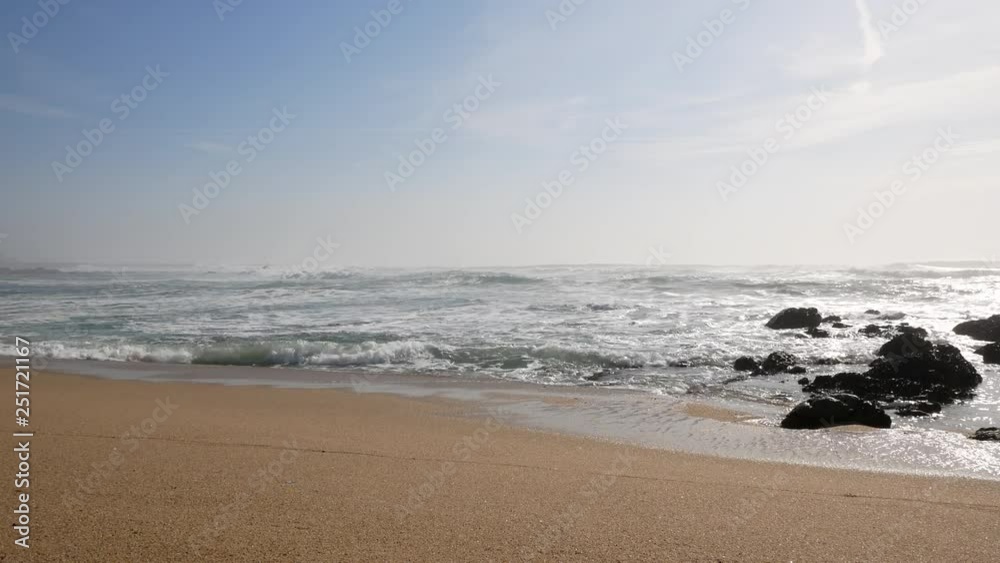 Wide angle view of white surf waves breaking on the sandy shore of the beach. Blue sky and rocks to right. Wide angle view of white surf waves breaking on the sandy shore of the beach. Blue sky and rocks to right.
