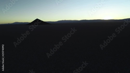 Aerial general view of silhuette of natural isolated pyamid in wide brown and white salar at sunset. Flying sideways near white floor towards cone. Golden hour. Abstract nature. Cono de Arita, Salta photo