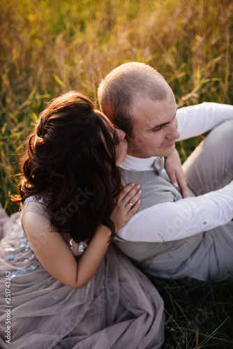 Wedding near the river in field at sunset with brown horse. Bride in light airy dress in color of dusty rose. Beige dress with sparkles. Light suit with bow tie. bride and groom embrace and kiss. © malysheva
