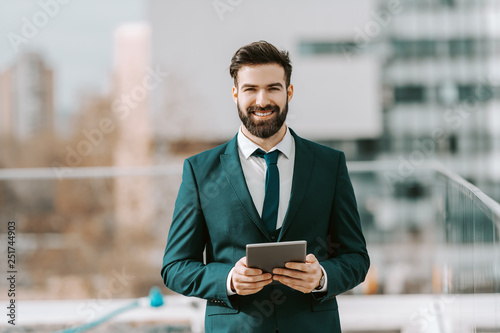 Smiling businessman in formal wear using tablet while standing on the rooftop. In background buildings and streets.
