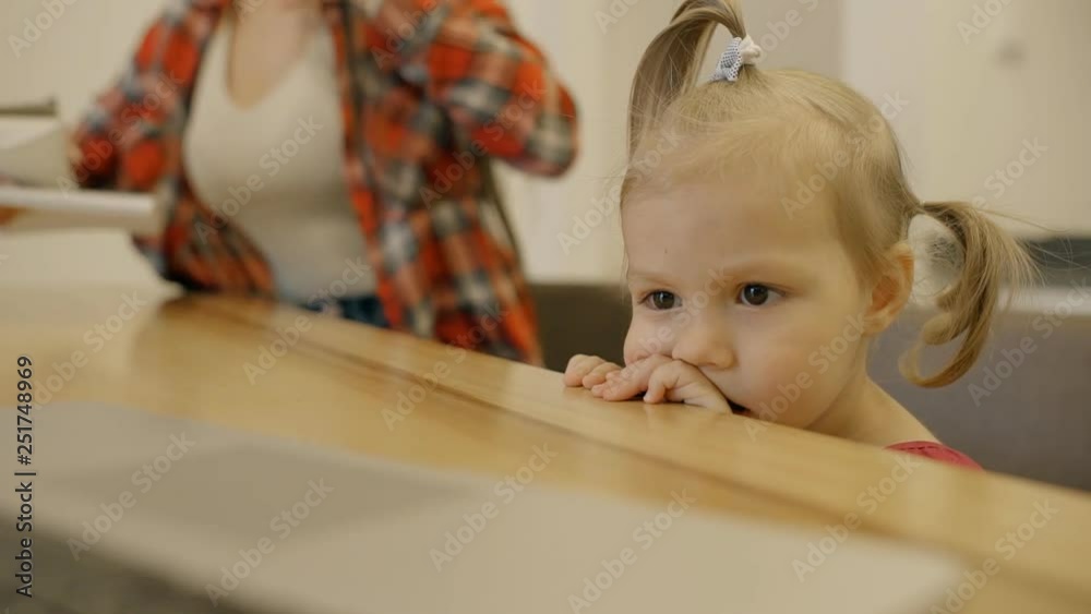 Little girl looks at monitor, woman sits down at table in home interior. Little girl looks at monitor, woman sits down at table in home interior.