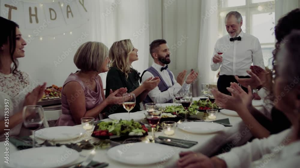 A big family sitting at a table on a indoor birthday party, a senior man giving a speech.
