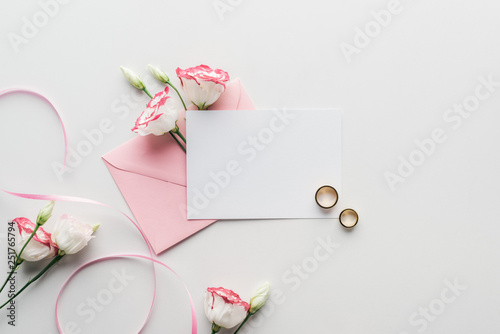 top view of empty card with pink envelope, flowers, silk ribbon and golden wedding rings on grey background