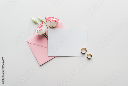 top view of empty card with pink envelope, flowers and golden wedding rings on grey background photo