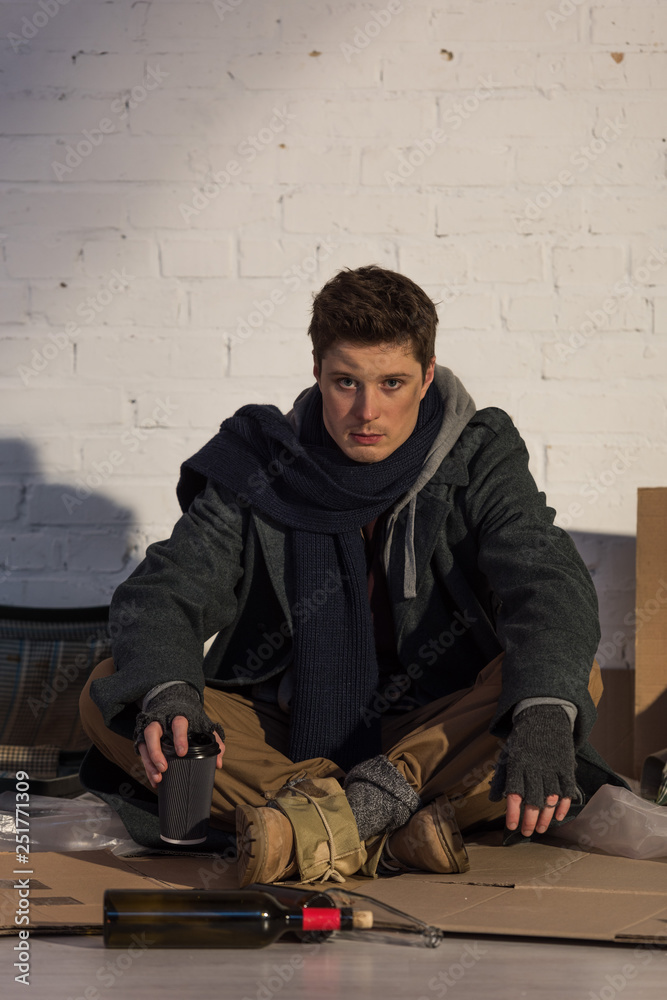 homeless man sitting on cardboard by white brick wall and holding paper cup homeless man sitting on cardboard by white brick wall and holding paper cup