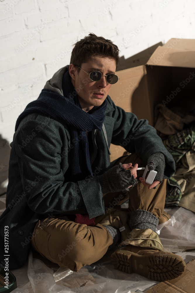 Obraz premium homeless man sitting by white brick wall and holding cigarette pack