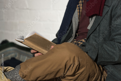 cropped view of homeless man reading book while sitting by white brick wall
