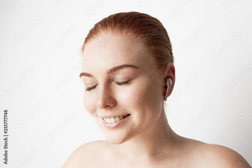 Portrait of charming redhead woman with romantic smile and closed eyes listening music with wireless earphone in ear over background Portrait of charming redhead woman with romantic smile and closed eyes listening music with wireless earphone in ear over background