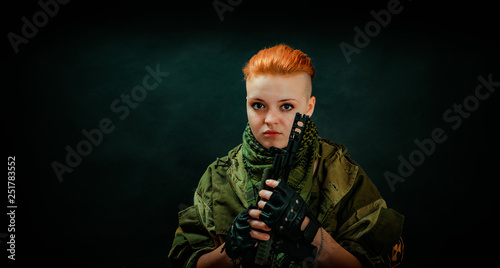 Woman with red hair in military uniform hold machine gun in darck room. Vertical photo