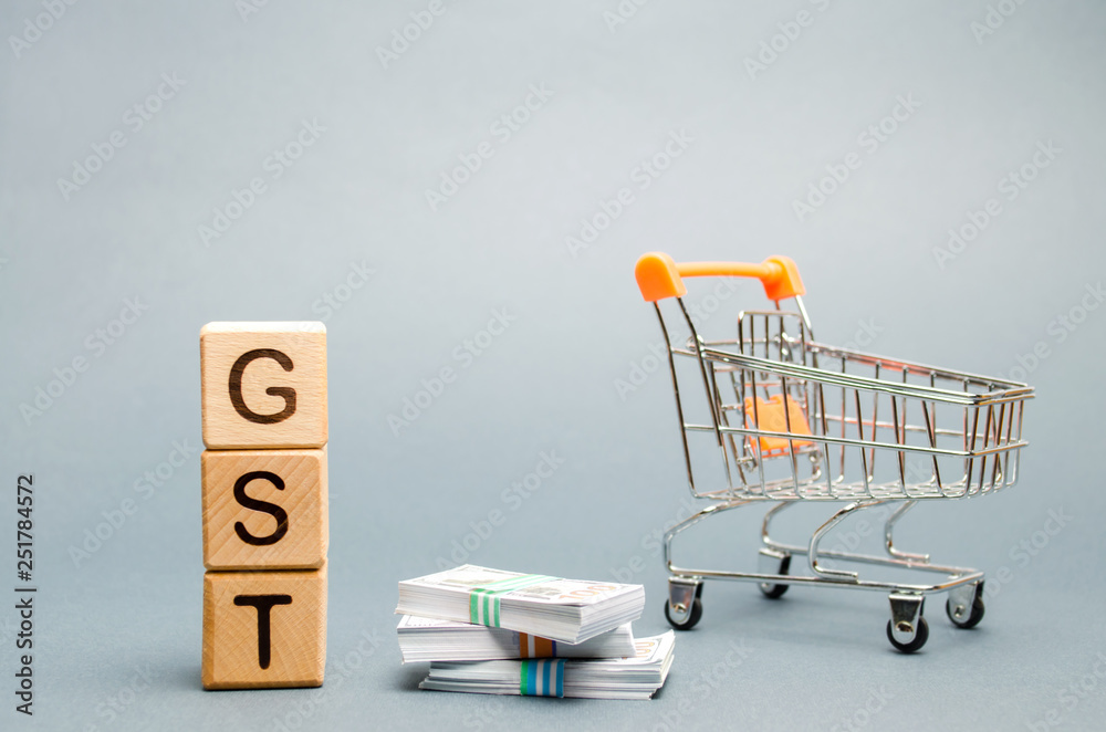 Wooden blocks with the word GST (Goods & Services Tax), money and a supermarket trolley. Tax, which is imposed on the sale of goods and services