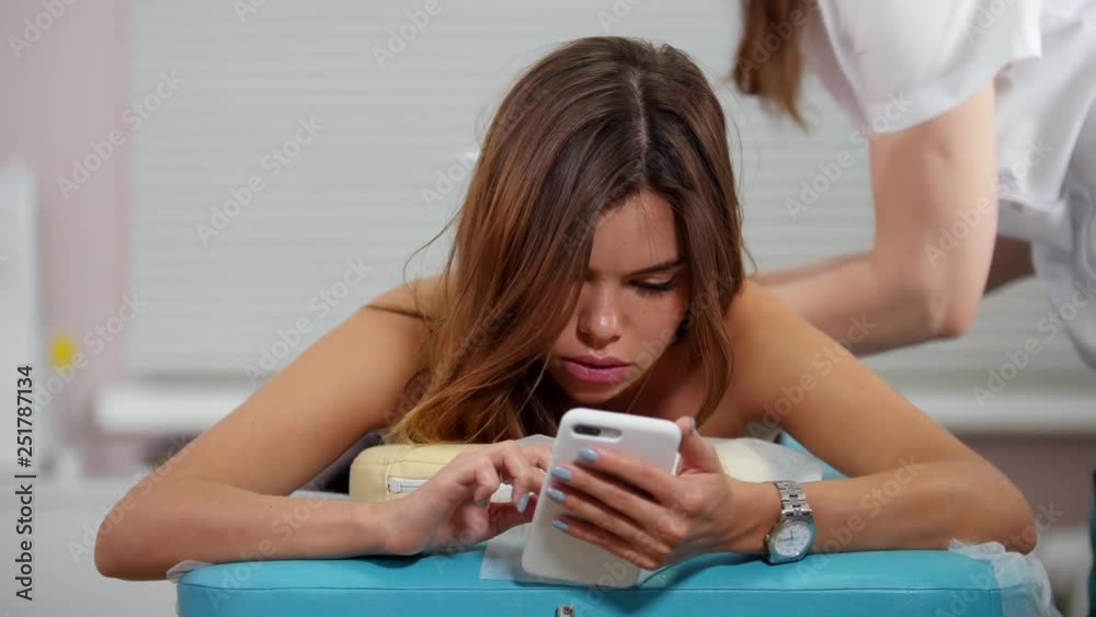 A young brunette woman receiving a relaxing back massage. Sitting in her phone and looking at the screen A young brunette woman receiving a relaxing back massage. Sitting in her phone and looking at the screen