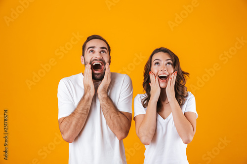 Image of happy people man and woman in basic clothing screaming in surprise and touching cheeks, isolated over yellow background