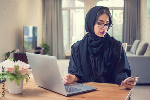 Beautiful Arab woman using smartphone and laptop while sitting in cozy apartment