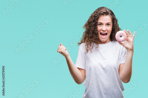 Young brunette girl eating donut over isolated background screaming proud and celebrating victory and success very excited, cheering emotion