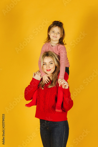 Happy little girl with her young mother on the yellow background. Daughter sitting on mom's neck. Smile
